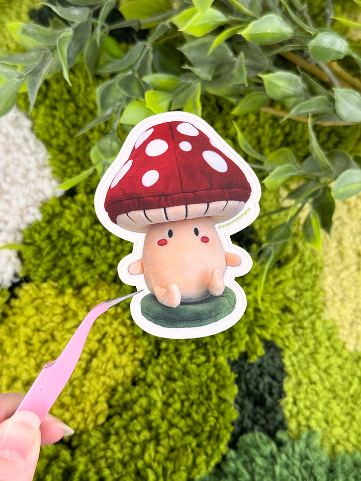 A hand holds tweezers gripping a cute mushroom sticker with a red cap and white spots, against a background of green plants and textured surface.
