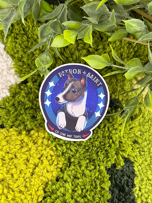 A sticker with an illustrated dog in a blue outfit, labeled Patron Saint of One Way Trips, is placed on green textured fabric beside artificial leaves.