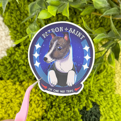 A hand holds tweezers grasping a sticker of a dog in a shirt with a starry background and the text PATRON SAINT OF ONE WAY TRIPS, surrounded by green foliage.