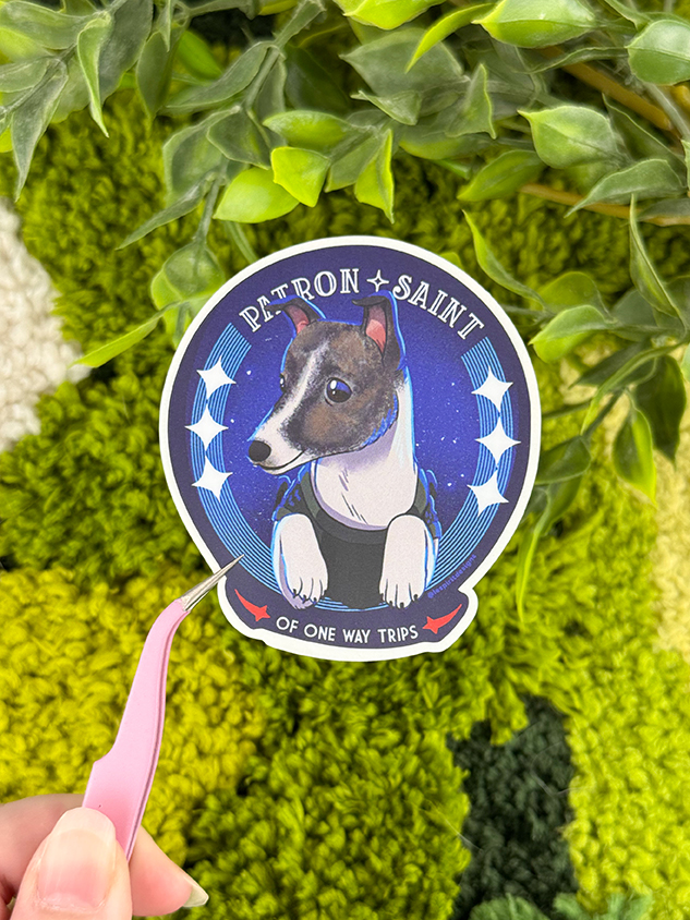 A hand holds tweezers grasping a sticker of a dog in a shirt with a starry background and the text PATRON SAINT OF ONE WAY TRIPS, surrounded by green foliage.
