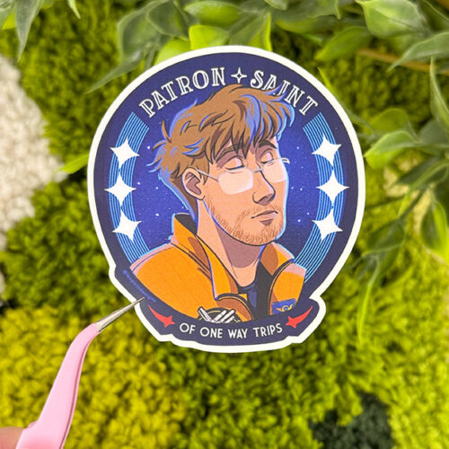 A hand holds a sticker showing an illustrated person with glasses and an orange jacket, surrounded by stars and a blue background. Text reads, Patron Saint of One Way Trips. Green plants are in the background.