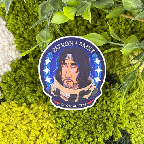 A sticker with an illustrated man and the text PATRON SAINT OF ONE WAY TRIPS, placed on textured green and white fabric, surrounded by green leaves.