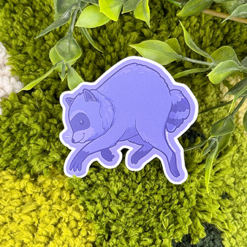 A purple raccoon sticker is placed on a textured, green and white fabric with leafy plant stems above it.