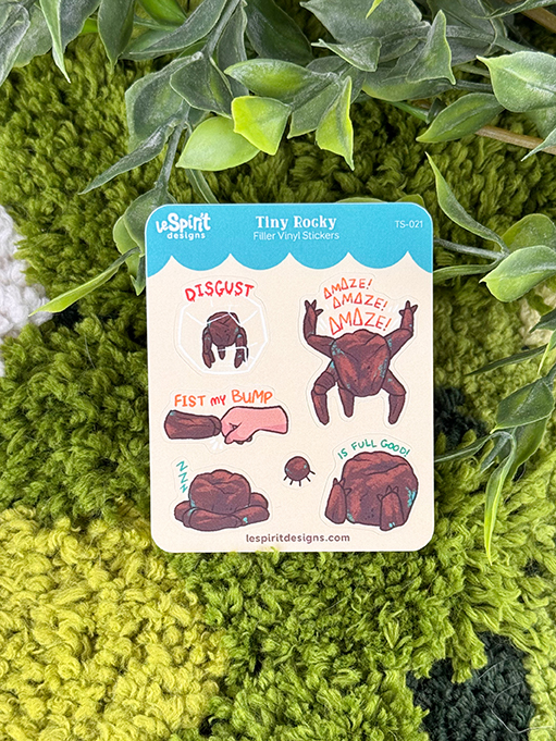 A Tiny Rocky ~ Tiny Filler Vinyl Sticker Sheet featuring cartoon rocks with expressive faces and phrases like “DISGUST,” “AMAZE! AMAZE! AMAZE!”, “FIST MY BUMP,” and “is full good!” is displayed on a green and white textured surface, accented by leafy plants overhead.
