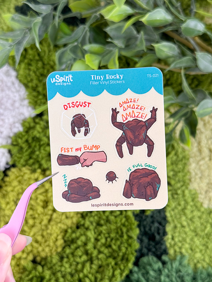 A hand holds the Tiny Rocky ~ Tiny Filler Vinyl Sticker Sheet, featuring cartoon rock characters expressing emotions like DISGUST, AMAZE!, Fist my bump, and IS FULL GOOD! Green plants are blurred in the background.