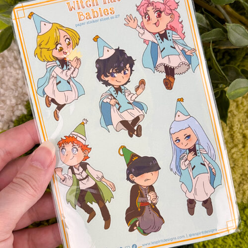 A hand holds a sheet of Witch Hat Babies paper stickers featuring six chibi-style characters in blue outfits and party hats, with green and yellow plants in the background.
