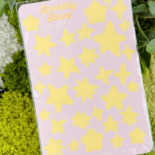 A sheet of yellow star-shaped stickers labeled Sparkly Stars on a light purple background, placed on green and white textured fabric with leaves around it.