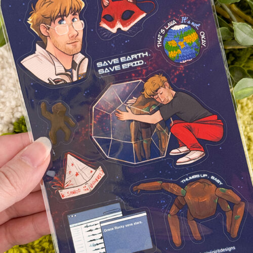 A hand holds a sticker sheet with illustrations: a man, a red mask, Earth with Thats a Lala its not okay, a man hugging a glass structure, a crab, a spaceship, and a computer screen with star data.