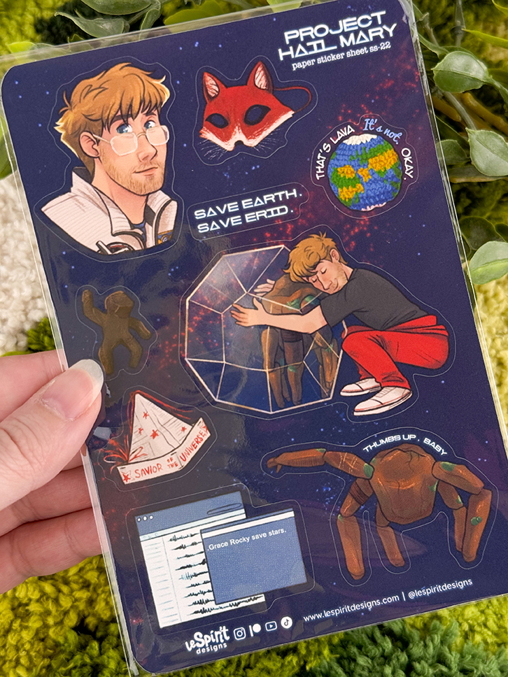 A hand holds a sticker sheet with illustrations: a man, a red mask, Earth with Thats a Lala its not okay, a man hugging a glass structure, a crab, a spaceship, and a computer screen with star data.
