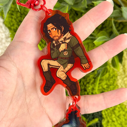 A hand holds a red acrylic keychain featuring a cartoon character in medieval clothing, with a small dangling piece showing a fish. Lush green plants are blurred in the background.