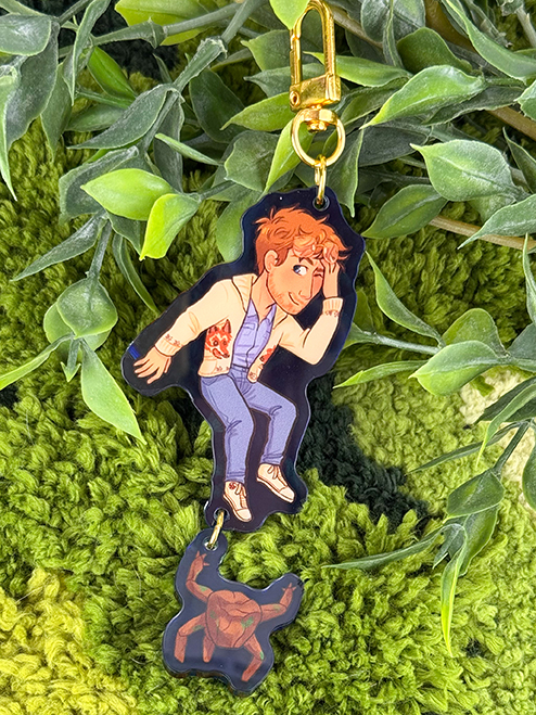 A keychain with a cartoon person in a denim jacket and a small turtle charm, displayed on green leafy and textured background.