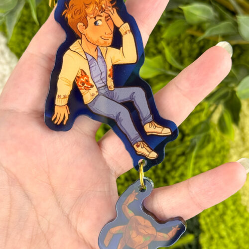 A hand holds a keychain showing an illustrated person in a tan jacket and jeans, with a small turtle charm hanging below. Green leafy plants are in the blurred background.