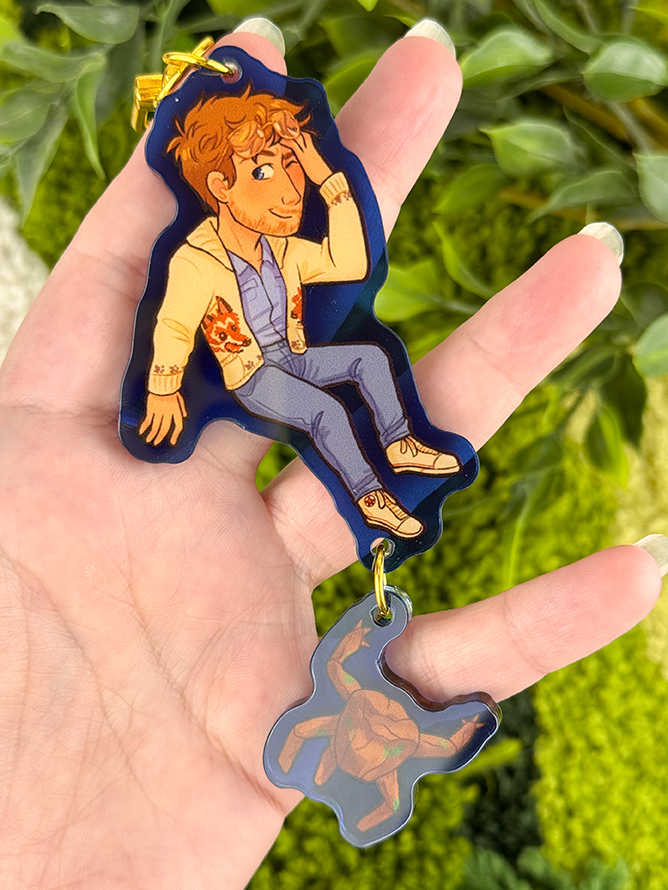 A hand holds a keychain showing an illustrated person in a tan jacket and jeans, with a small turtle charm hanging below. Green leafy plants are in the blurred background.
