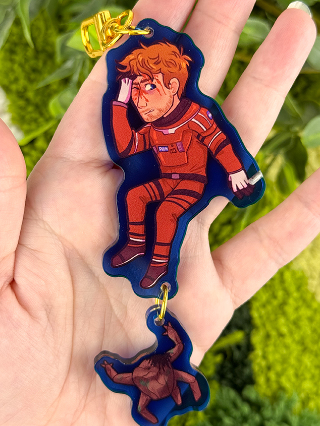 A hand holds a keychain featuring an illustrated astronaut in a red suit, saluting, and a small dinosaur charm attached below. Green foliage is blurred in the background.