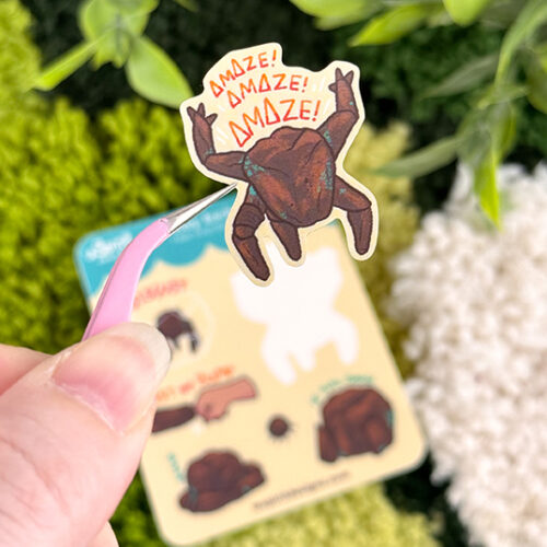 A hand holds tweezers gripping a sticker from the Tiny Rocky sticker sheet, with greenery and a blurred illustration card in the background.