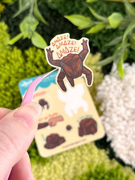 A hand holds tweezers gripping a sticker from the Tiny Rocky sticker sheet, with greenery and a blurred illustration card in the background.