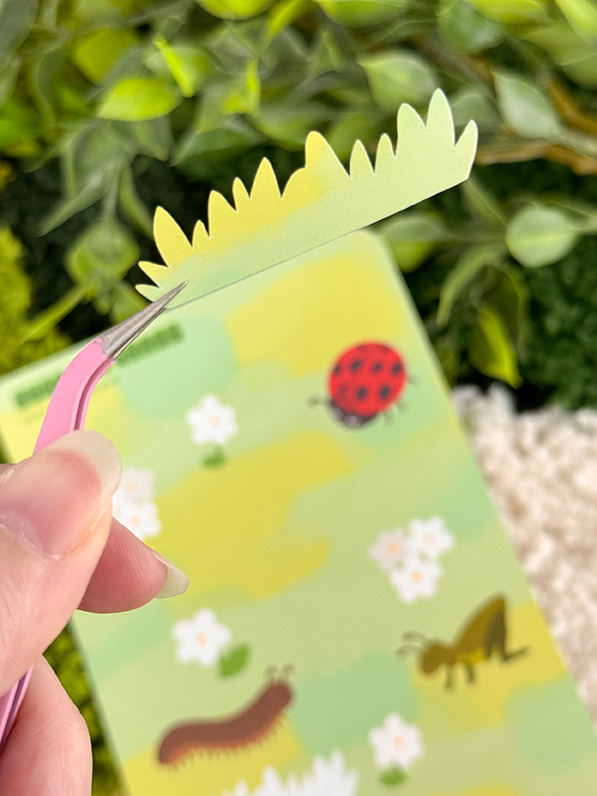 A hand holds tweezers gripping a grass-shaped sticker over a nature-themed background with flowers, a ladybug, a caterpillar, and a praying mantis, surrounded by green plants.