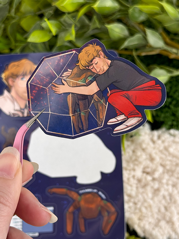 A hand holds a sticker of a person in red pants hugging a transparent, geometric shape with another figure inside. Green leaves and more stickers appear blurred in the background.