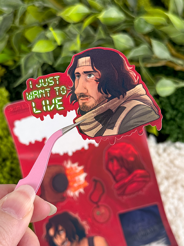 A hand holds tweezers with a sticker of a serious man with long dark hair, a headband, and scarf. Neon green text reads I JUST WANT TO LIVE. Blurred greenery and more stickers are in the background.