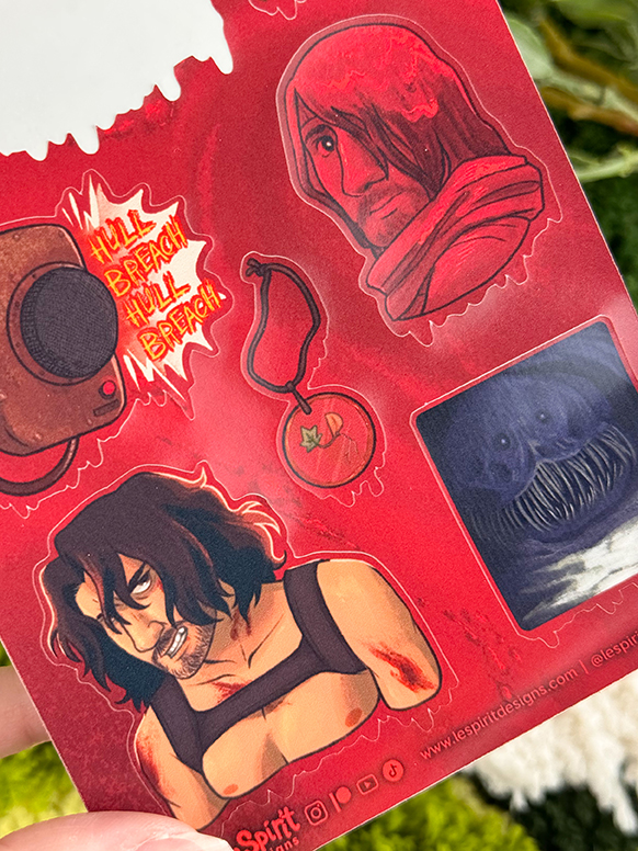 A sheet of red stickers featuring illustrated characters, a blood-splattered HILL BEACH! speech bubble, a tomato keychain, and a creepy, toothy monster face, all held by a hand.