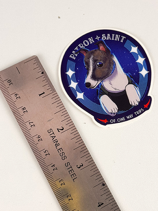 A Patron Saint of One Way Trips sticker with a dog in a spacesuit, next to a stainless steel ruler for scale.