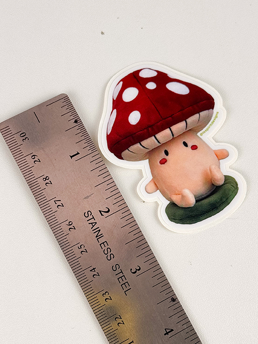 A sticker of a cute cartoon mushroom character beside a stainless steel ruler showing it is about 2 inches tall.