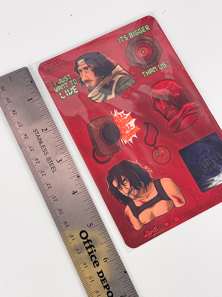 A red sticker sheet with illustrated characters, text, and objects from the game Iron Lung, next to a stainless steel ruler marked Office Depot..
