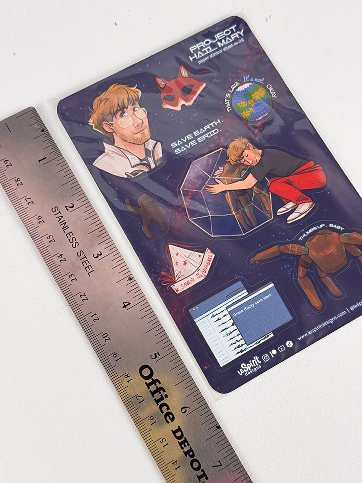 A sheet of Project Hail Mary themed stickers lies next to a 12-inch stainless steel ruler. The stickers include a person, a rock, a spaceship, Earth, and other items from the story.