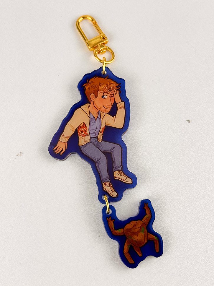A keychain with a cartoon character waving and a small creature below, connected by metal links. The keychain has a gold clasp and features bright, colorful artwork on a white background.