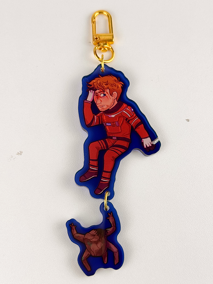 A keychain with a cartoon astronaut in a red suit above a small, brown, furry creature. Both charms are connected by a gold clasp and chain, displayed on a white background.
