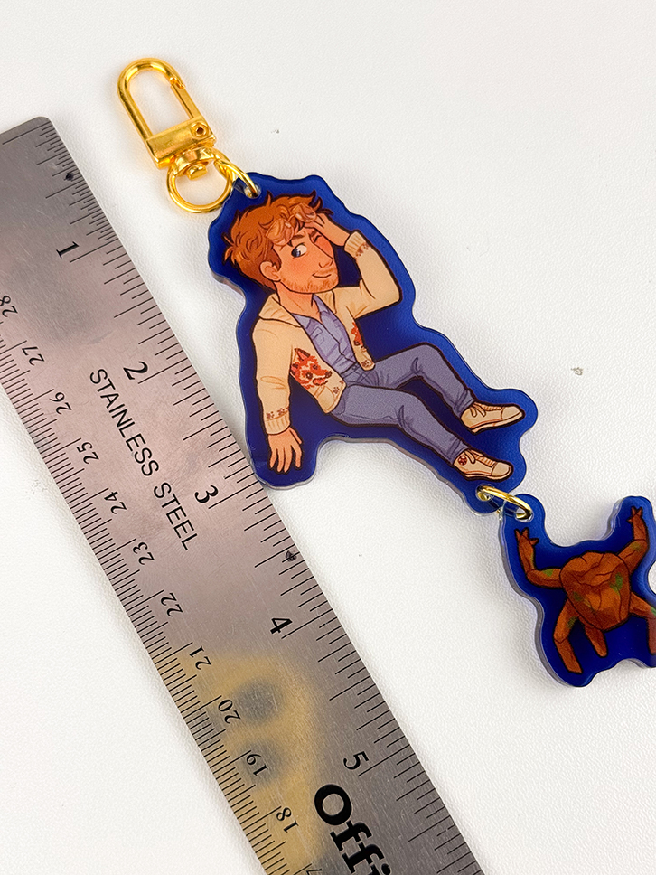 A stainless steel ruler is next to a keychain showing a cartoon person in casual clothes and a smaller turtle charm. The keychain has a gold clasp and is about 9 cm long.