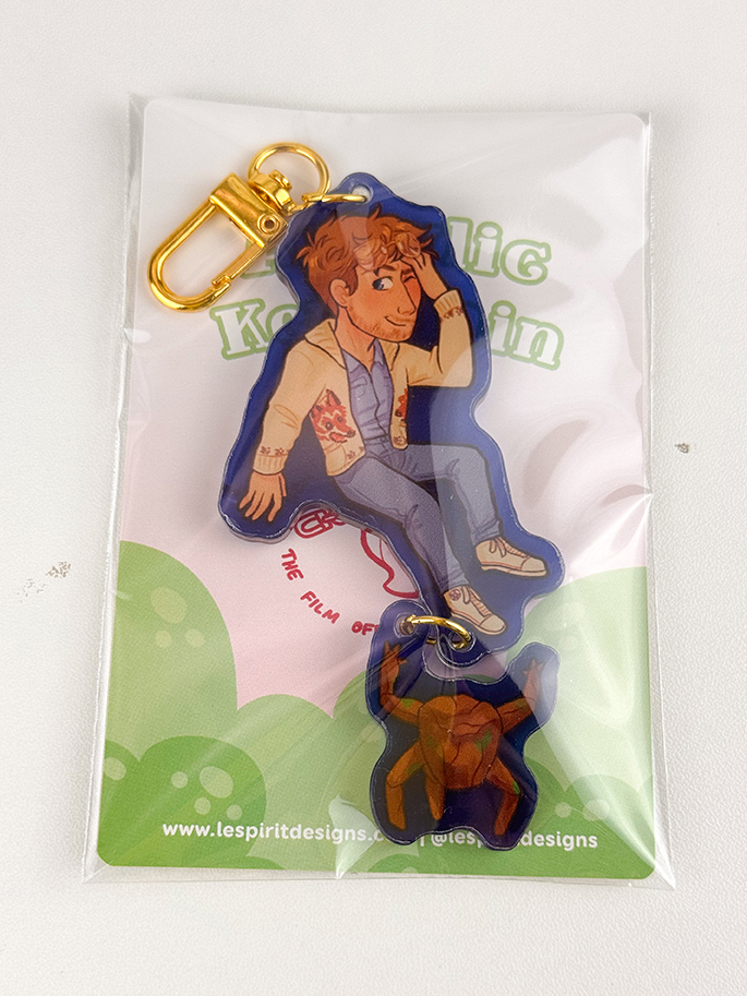 A keychain with a cartoon character in a blue jacket and jeans, sitting and smiling, attached to a gold clasp. The keychain is packaged on a green and white backing card.