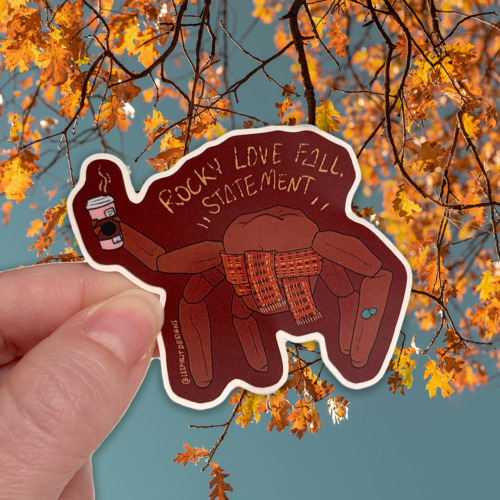 A hand holds a Witch Hat Babies ~ Paper Sticker Sheet (Copy) sticker of a brown crab wearing a plaid scarf and holding a coffee cup, with text reading Rocky love fall.